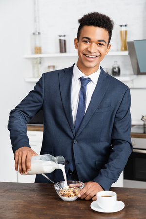 happy african american businessman pouring fresh milk in bowl with corn flakesの写真素材