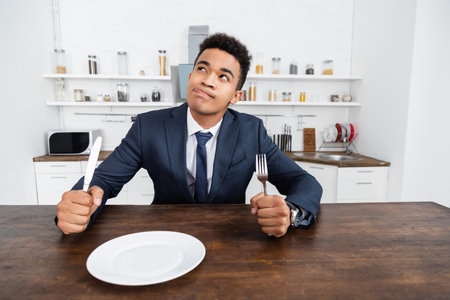 dreamy african american man holding cutlery while thinking and sticking out tongue