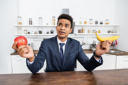 puzzled african american businessman choosing between apple and bananaの写真素材