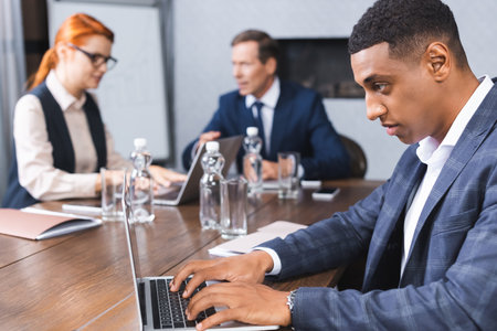 Concentrated african american businessman typing on laptop at workplace with blurred colleagues on backgroundの写真素材