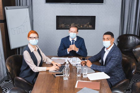 Multicultural businesspeople in medical masks looking at camera while sitting at workplace with digital devices in meeting roomの写真素材