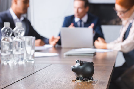 Small black piggy bank with coins on table with blurred multicultural businesspeople on backgroundの写真素材
