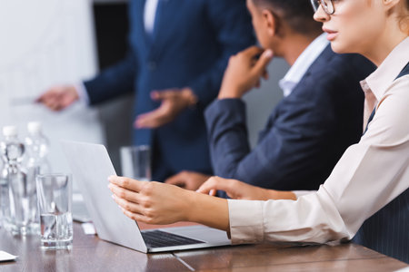 Cropped view of businesswoman using laptop while sitting at workplace with blurred multicultural colleagues on backgroundの写真素材
