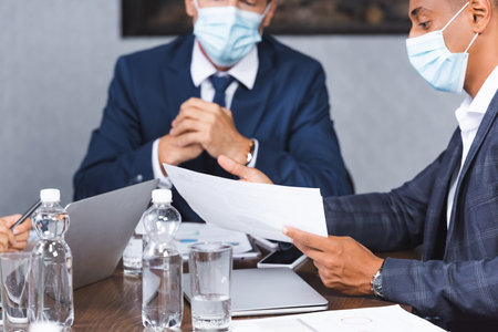 African american businessman in medical mask showing papers while sitting at workplace with blurred colleague on backgroundの写真素材