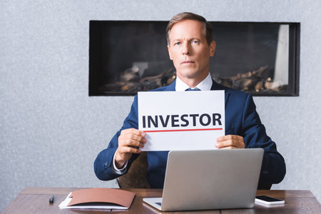 Serious businessman looking at camera and holding sign with investor lettering at workplace with digital devices in meeting roomの写真素材