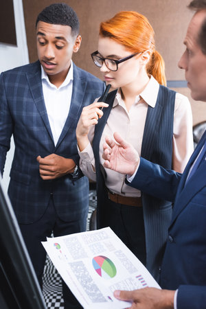 Thoughtful redhead businesswoman standing near multicultural colleagues talking and gesturing on blurred foregroundの写真素材