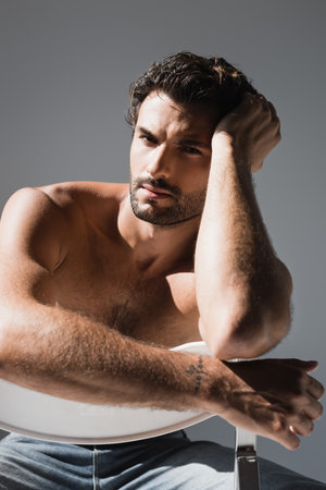 Shirtless man in jeans looking at camera while sitting on chair isolated on grayの写真素材
