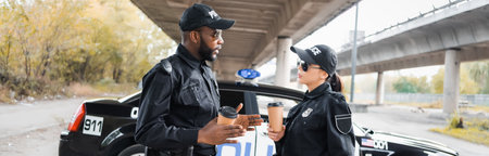 multicultural police officers with paper cups talking near patrol car on blurred background on urban...