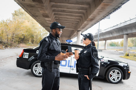multicultural police officers with paper cups talking near patrol car on blurred background on urban...