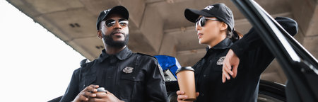multicultural police officers with paper cups talking while leaning on patrol car on blurred...
