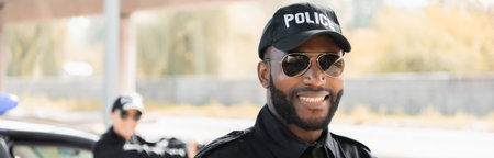 portrait of happy african american policeman looking at camera with blurred colleague on background outdoors, bannerの写真素材