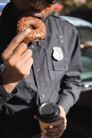 cropped view of african american policeman with paper cup eating donut on blurred background...