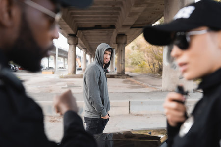 dissatisfied hooded offender looking at blurred multicultural police officers on foreground on urban...