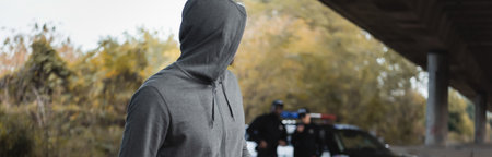 hooded offender looking away with blurred multicultural police officers on background outdoors,...