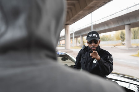 serious african american policeman pointing with finger at blurred hooded offender on foreground on urban streetの写真素材