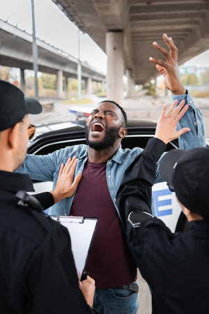 back view of police officers calming despaired african american man near patrol car on blurred background on urban streetの写真素材