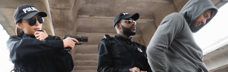 african american policeman arresting hooded offender near colleague aiming by pistol outdoors,...
