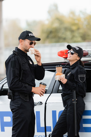 Smiling policewoman in sunglasses holding burger and coffee to go near colleague eating and car...