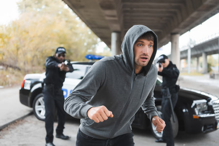 scared hooded offender running from multicultural police officers aiming by gun on blurred background outdoorsの写真素材