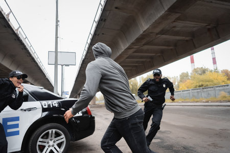 hooded offender running from multicultural police officers near patrol car on urban streetの写真素材