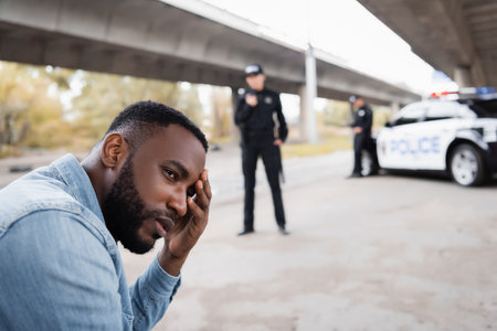 Upset african american victim looking away near police officers and car on blurred background on urban streetの写真素材