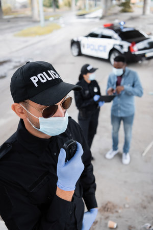 Policeman in latex gloves and medical mask using walkie talkie while colleague talking with african...