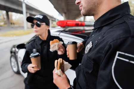 Policeman holding burger and coffee to go near colleague and car on blurred background on urban...