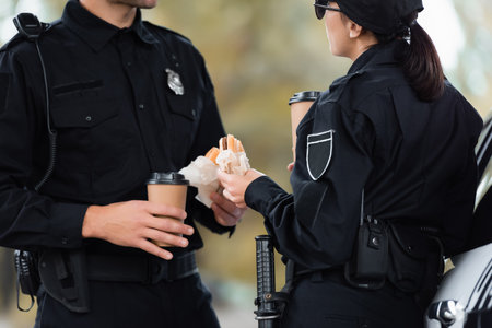 Policewoman in uniform holding burger and takeaway coffee near colleague and car on blurred foregroundの写真素材