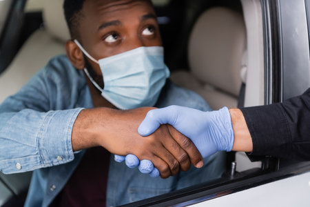 Police officer in latex glove handshaking with african american driver in medical mask on blurred background in carの写真素材