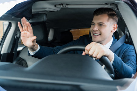Cheerful businessman waving hand while driving car on blurred foreground
