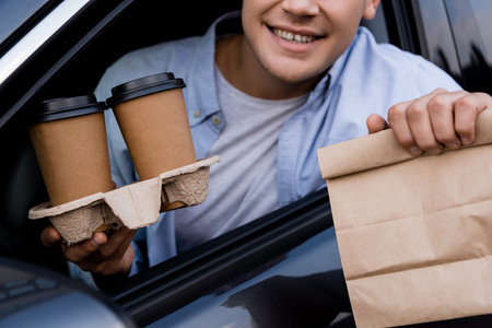 cropped view of smiling man holding paper bag and coffee to go in car, blurred foregroundの写真素材