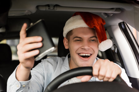 drunk, cheerful man in santa hat holding flask with alcohol while driving car, blurred foregroundの写真素材