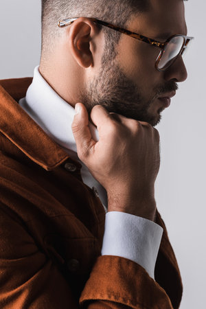 Young man in eyeglasses adjusting collar of white turtleneck isolated on gray