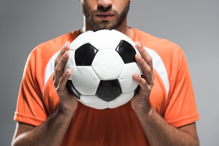 Cropped view of football in hands of bearded sportsman on blurred background isolated on gray