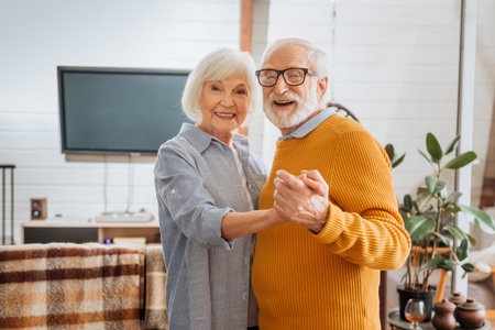 happy elderly couple looking at camera while dancing at home on blurred background
