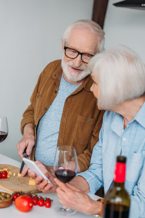 smiling senior husband looking at wife while cutting cheese on chopping board near vegetables on blurred foregroundの写真素材