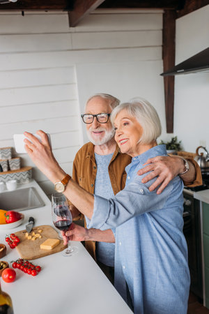 smiling elderly couple with wine glasses hugging while taking selfie near table with food in kitchenの写真素材