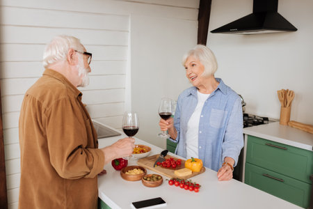 smiling senior man with wine glass looking at husband near table with food in kitchenの写真素材