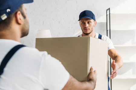 young worker in uniform taking carton box near indian coworker on blurred foregroundの写真素材
