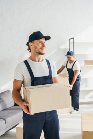 young worker in uniform holding carton box near indian coworkerの写真素材