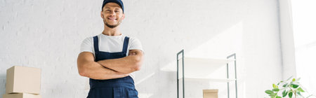 happy mover in uniform and cap standing with crossed arms in apartment, bannerの写真素材