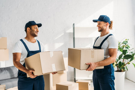 happy multiethnic movers in caps and uniform carrying boxes in apartmentの写真素材