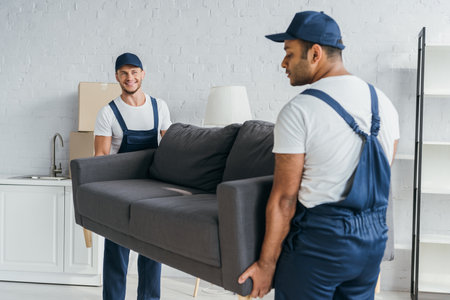 young mover in uniform smiling while carrying couch with indian colleague on blurred foregroundの写真素材
