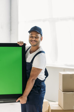 happy indian mover in uniform carrying plasma tv with green screen in apartmentの写真素材