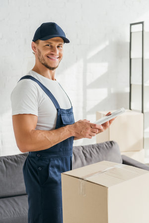 happy mover in uniform holding digital tablet in apartmentの写真素材