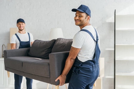 young indian mover in uniform smiling while carrying couch with colleague on blurred backgroundの写真素材
