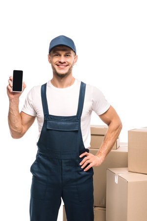 cheerful mover in uniform holding smartphone with blank screen and standing with hand on hip near carton boxes isolated on whiteの写真素材
