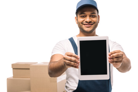 happy indian mover in overalls holding digital tablet with blank screen near carton boxes on blurred background isolated on whiteの写真素材