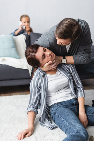Abusive husband choking wife with closed eyes while kid talking on smartphone on blurred backgroundの写真素材