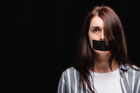 Victim of domestic violence with adhesive tape on mouth looking at camera isolated on black